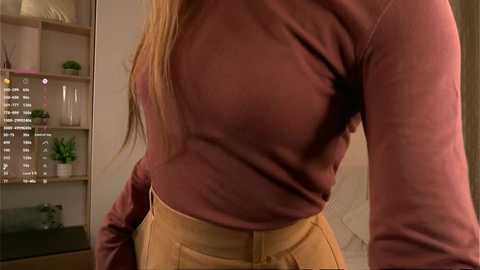 Media: Video of a woman with long blonde hair, wearing a fitted, long-sleeved, mauve turtleneck and high-waisted mustard yellow skirt, standing indoors next to a bookshelf with potted plants.