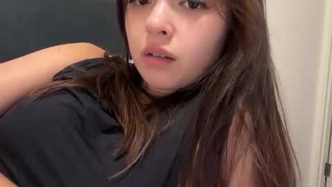 Video of a young woman with long brown hair, fair skin, and brown eyes, wearing a black sleeveless top, indoors with blurred background.