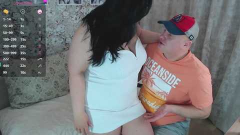 Video of a plus-size woman with long black hair in a white dress, being held by a bald, overweight man in an orange shirt and cap.