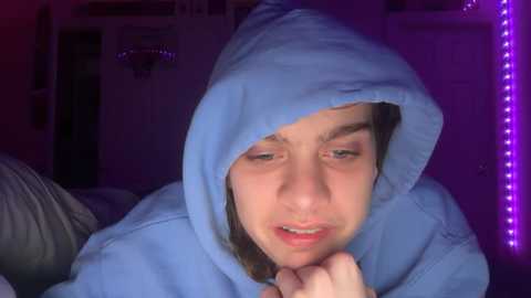 Video of a young woman with fair skin and light brown hair, wearing a blue hoodie with hood up, looking contemplative with mouth slightly open, in a dimly lit room with purple LED lights on the right.