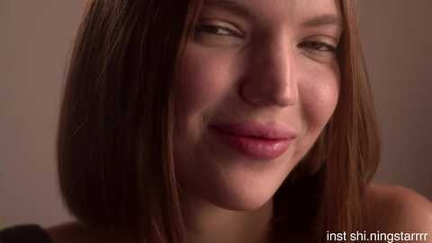 Close-up video of a young woman with straight, shoulder-length brown hair, fair skin, and a subtle smile. She wears minimal makeup and a black top. The background is blurred and neutral.