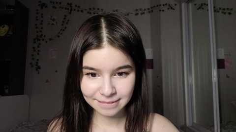 Video of a young woman with straight, shoulder-length dark brown hair and fair skin, smiling softly. Background features a dimly lit room with string lights, a dark wall, and a white mirror.