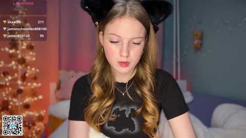A video of a young Caucasian girl with long, wavy blonde hair, wearing black cat ears and a black t-shirt, sitting in a softly lit room with a Christmas tree and wall decorations.