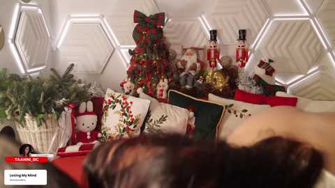 Video of a cozy, festive living room adorned with a Christmas tree, plush toys, and holiday-themed cushions on a white couch. Hexagonal white lights illuminate the background. A person's head is partially visible in the foreground.