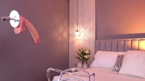 Video of a modern bedroom with a grey wall, white bed, white pillows, and a metallic bedside table. A realistic, erect penis is mounted on a metal arm, pointing towards the bed.
