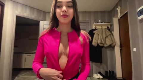 Video of a young woman with long brown hair, wearing a vibrant pink, low-cut top, standing in a modestly furnished room with beige walls, a wooden door, and hanging coats.