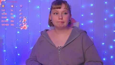 Video of a young Caucasian woman with short brown hair, light skin, and a slight build, wearing a gray hoodie. She stands against a backdrop of blue fairy lights and a calendar with cartoon characters.