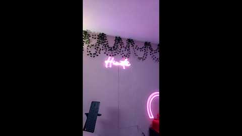 A video of a dimly lit room with a pink neon sign spelling \"love\" above a white wall adorned with ivy. A blue chair is visible on the left, and a pink neon lightbulb hangs on the right.