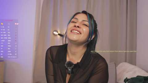 Media: Video of a smiling, light-skinned woman with blue-streaked hair in a sheer black blouse, sitting in a dimly lit room with a computer screen displaying chat messages.
