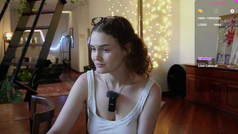 Video of a young woman with curly brown hair, wearing a white tank top, seated indoors with a microphone, surrounded by cozy, warmly lit room with wooden furniture and Christmas decorations.