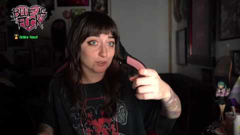 Video of a pale, brunette woman with bangs, wearing a black graphic tee, adjusting a pink headset mic. Background shows dark room, clutter, and a toy figure.