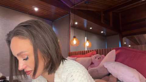 Media: Video of a young woman with straight, shoulder-length brown hair, wearing a white knit sweater, sitting on a bed with plush pink and white teddy bears. The room features warm wooden ceiling beams and soft lighting.