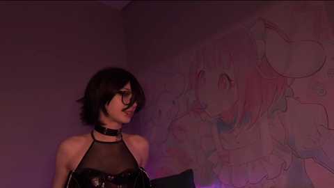 Video of a slender, pale-skinned woman with short black hair, wearing a black leather choker and halter top, standing against a wall with a pink, anime-style drawing.