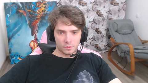 Video of a young man with short brown hair wearing a black t-shirt, headphones, and a headset, sitting in a pink gaming chair with a vibrant blue and orange mural behind him.