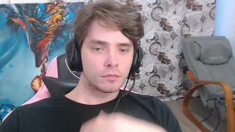 Video of a young Caucasian man with brown hair, wearing a black T-shirt and headphones, sitting in a gaming chair, with a vibrant dragon and motorcycle-themed backdrop.