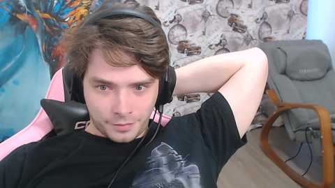 Video of a young man with short brown hair, wearing headphones and a black T-shirt, seated in a pink gaming chair, surrounded by a gray sofa and wall art.