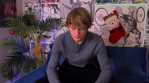 Video of a woman with short blonde hair in a blue chair, wearing a gray sweater, in a room with a cartoonish mural, potted plants, and a Christmas stocking.