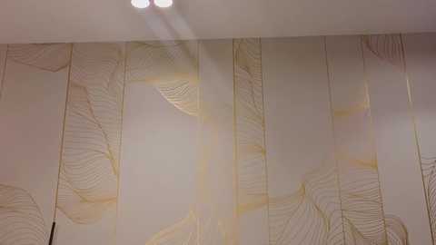 Media: Video of a modern, beige and gold-patterned wallpaper with abstract leaf designs, applied to a wall. The design features vertical, delicate leaf lines in varying shades of gold and beige, creating a textured, elegant aesthetic.