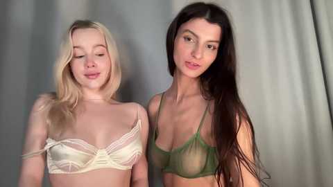 Video of two young women, one with fair skin and long blonde hair, wearing a sheer white bra, the other with olive skin and long dark hair, wearing a green bralette. Both have a serene expression against a muted, light-gray background.