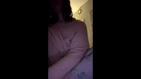 Video of a person with shoulder-length dark hair wearing a light-colored, possibly pink, cardigan, partially obscured by shadows. Background features a bed with floral bedding and a wall with a framed picture.