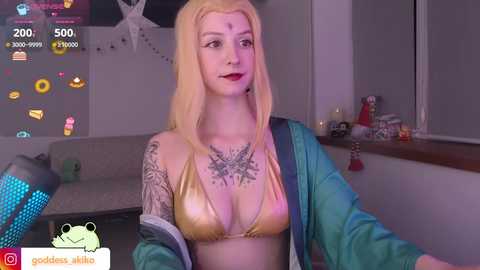 Media: Video of a pale-skinned, blonde-haired woman with a large tattoo on her chest, wearing a metallic gold bikini top and blue jacket, in a dimly-lit room with a TV screen displaying chat messages.
