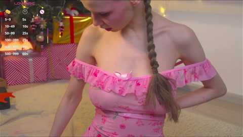 Video of a slender, fair-skinned woman with long blonde braided hair, wearing a pink, off-the-shoulder dress with floral patterns. She stands indoors near a decorated Christmas tree and presents, with a video game overlay in the top left corner.