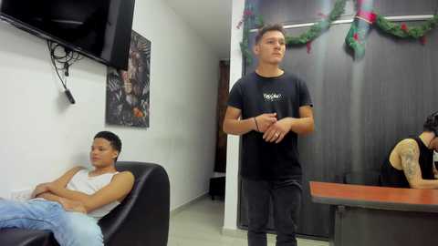 Video of a muscular young man in a black t-shirt and gray sweatpants, standing in a minimalist, white-walled room with a black leather chair and a small table.