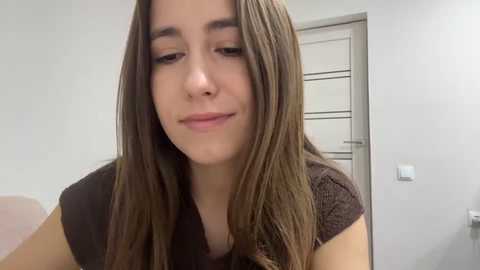 Video of a young woman with long, straight brown hair, wearing a dark brown top, smiling slightly in a modern, minimalist room with white walls and a closed door.