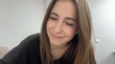 Video of a young Caucasian woman with straight brown hair, fair skin, and closed eyes, smiling gently. She wears a black top, and the background shows a plain beige wall and a white light switch.