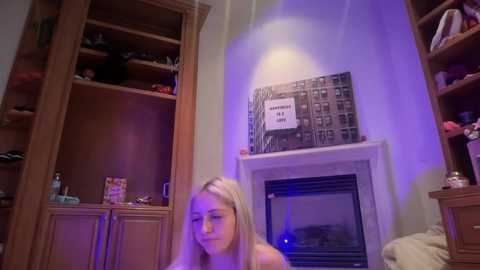 Video of a young blonde woman with long hair, sitting on a bed, in a dimly lit room with wooden furniture, bookshelves, and a framed poster.