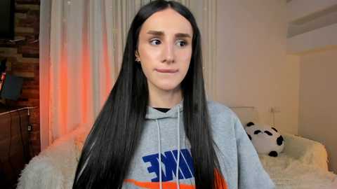 Media: Video of a young person with long black hair, wearing a grey hoodie with blue and orange text, in a cozy bedroom with a white bed, fluffy blanket, and a soccer ball.