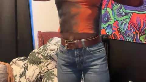 Video of a person in a tight orange tank top, blue jeans, and a brown belt, standing in a bedroom with a colorful, patterned bedspread and a vibrant, floral-patterned wall hanging.