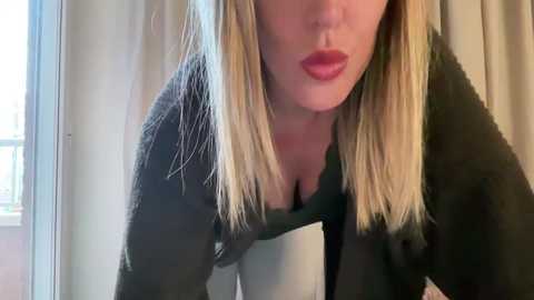 Media: Video of a blonde woman with light skin and red lipstick, wearing a black sweater, leaning against a window with sheer curtains, creating a soft, natural light effect.