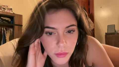 A video of a young woman with long brown hair, wearing a light-colored top, looking contemplative. Background shows a cluttered room with wooden furniture and a bookshelf.