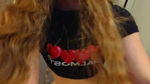 A video of a fair-skinned woman with long, wavy, light brown hair wearing a black crop top with a red heart necklace and a bold red heart design on her chest.