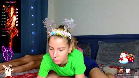 Media: Video of a young girl with blonde hair wearing a festive headband, lying on a bed with a Christmas-themed digital fireplace and a Santa Claus plush toy in the background.