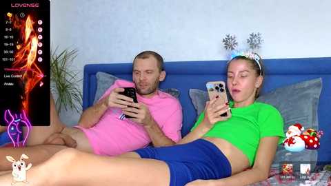 Media: Video of a man in a pink bodysuit and woman in a green crop top and blue shorts on a blue bed, watching a \"Love Island\" TV show.