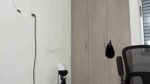 Video of a minimalist office corner featuring a white wall, a wooden cabinet with a black hook, a black mesh chair, and a white and black power strip.