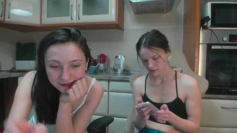 Video of two young women with fair skin and dark hair, one in a white tank top, the other in a black bra, sitting in a modern kitchen with white cabinets and stainless steel appliances.