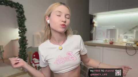 Media: Video of a young blonde woman with fair skin, wearing a white crop top with pink text and a gold pendant necklace, making a kissy face in a modern kitchen.