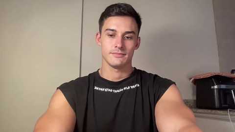 Video of a young man with short dark hair, wearing a black sleeveless shirt with white text, standing in a kitchen with beige cabinets and a black coffee maker.