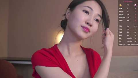 Video of an East Asian woman with fair skin and dark hair, wearing a red dress, adjusting her hair while looking at a digital calendar in a dimly lit room.