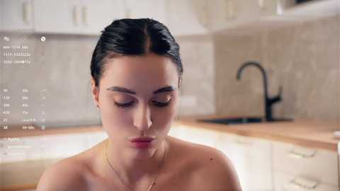 Media: Video of a topless woman with dark hair, fair skin, and closed eyes, standing in a modern kitchen with light cabinets and a black faucet.