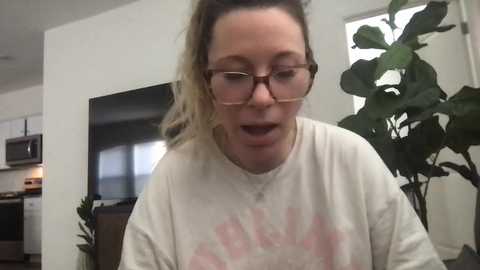 Media: Video of a light-skinned woman with shoulder-length blonde hair, wearing glasses and a white t-shirt with a pink graphic, smiling, in a modern kitchen with white walls, a large potted plant, and a microwave.