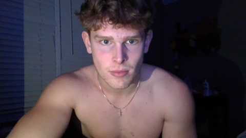 Media: Video of a shirtless young Caucasian man with curly brown hair, light skin, and a thin build, wearing a necklace, indoors with dim lighting.