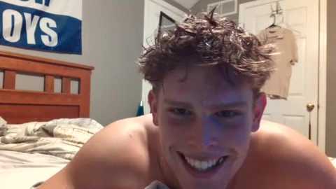 Media: Video of a shirtless, young, muscular man with curly brown hair, smiling, lying on a bed with a wooden headboard, surrounded by messy sheets and a door with a jacket hanging.