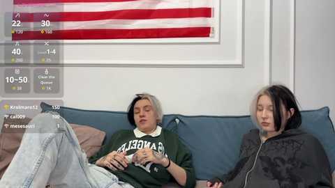 Video of two young women, one with short, dyed hair, in a casual setting, lying on a couch with a U.S. flag in the background.