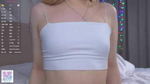 Media: Video of a fair-skinned, blonde woman with a small tattoo on her arm, wearing a white, spaghetti-strap crop top, sitting on a bed with white sheets, in a dimly lit bedroom with multicolored fairy lights.