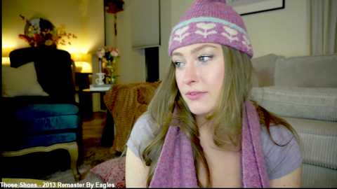 A video depicts a Caucasian woman with long brown hair, wearing a pink knit hat and scarf, sitting indoors, looking contemplative. The background features a cozy living room with warm lighting.