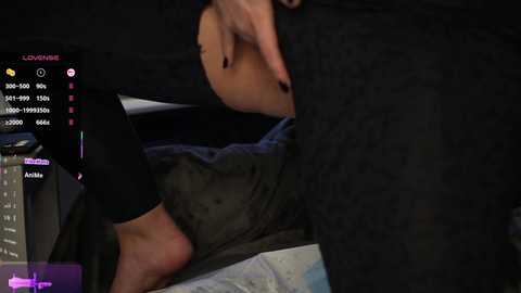 Media: Video of a woman with fair skin, black nail polish, and dark hair, sitting on a bed, wearing a black top and gray pants, with a gaming controller in her right hand.